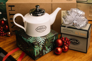 A white tea pot on top of a tree-themed gift wrapped box next to a box of tea bags with a ribbon tied in a bow on it with red ornament balls between them.